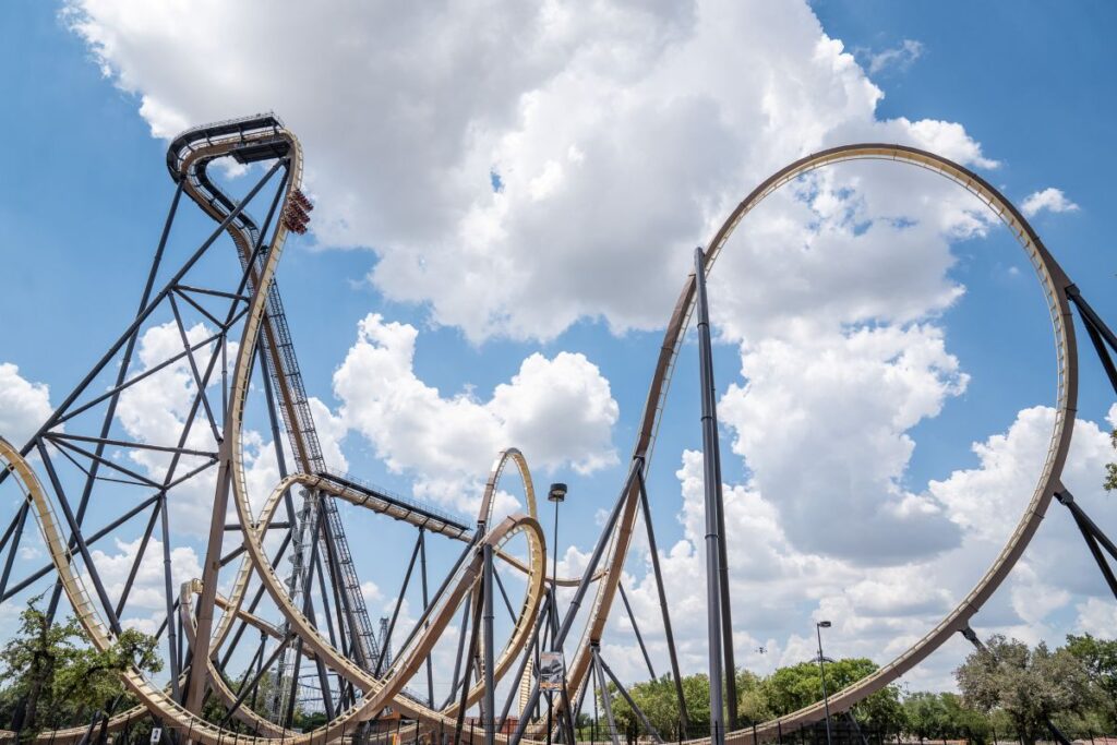 A wide view of a massive steel roller coaster layout featuring a towering vertical drop and large loops. The complex network of black support pillars and tan track stands out against a bright, cloudy sky.