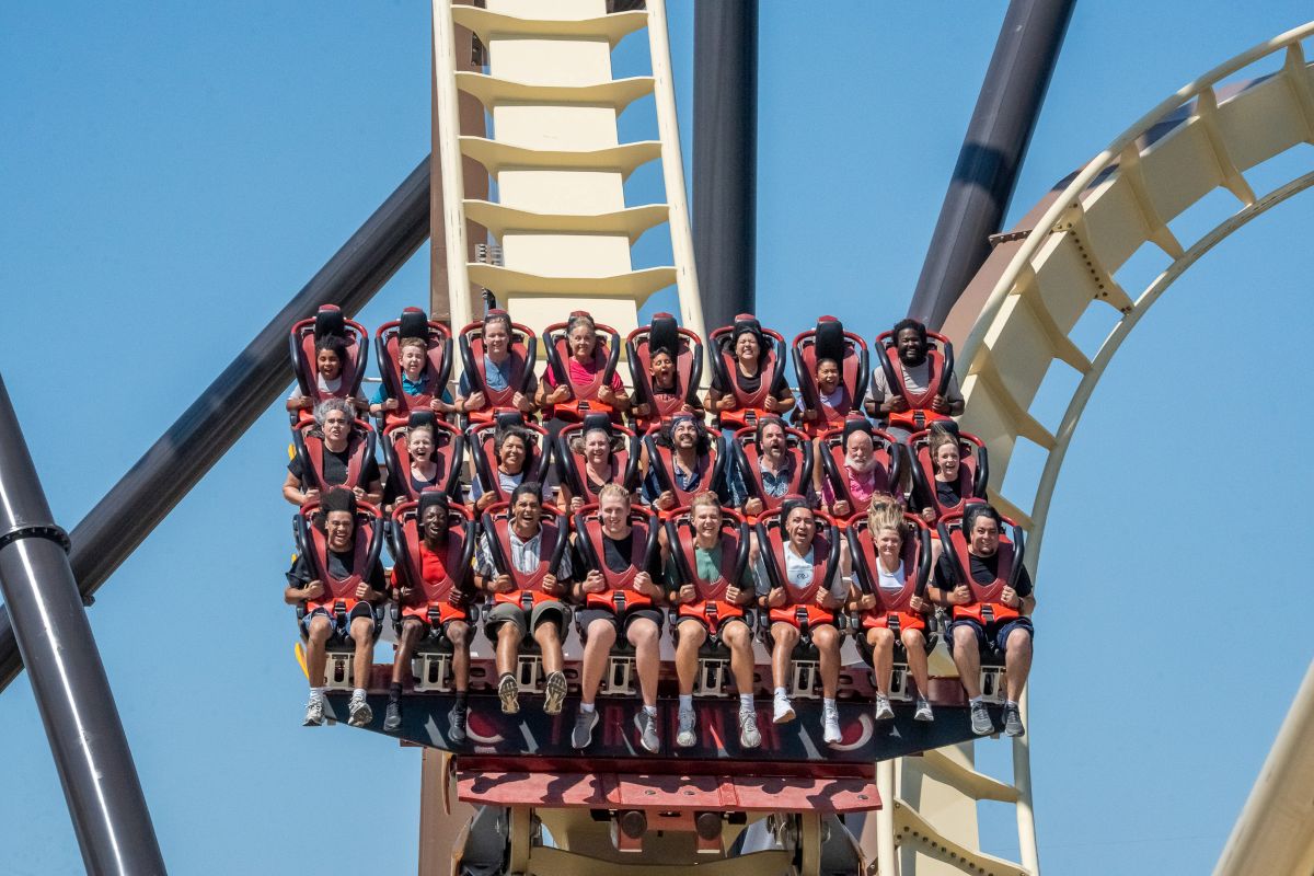 A front-facing view of a wide, multi-row roller coaster train carrying diverse riders down a drop. The passengers exhibit a mix of thrilled, smiling, and shouting expressions as they descend along the cream-colored track.