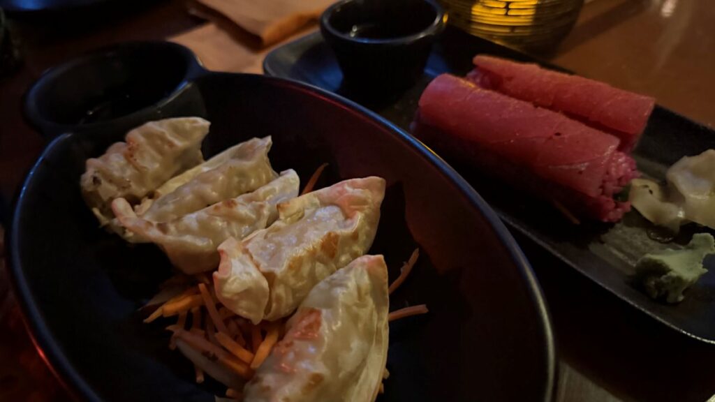 A close-up of pan-fried dumplings served over shredded carrots alongside a fresh tuna roll sushi dish under warm, dim restaurant lighting.