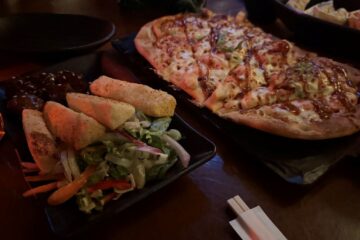 A flatbread pizza drizzled with sauce alongside a plate of seasoned potato wedges and a small side salad on a dark restaurant table.