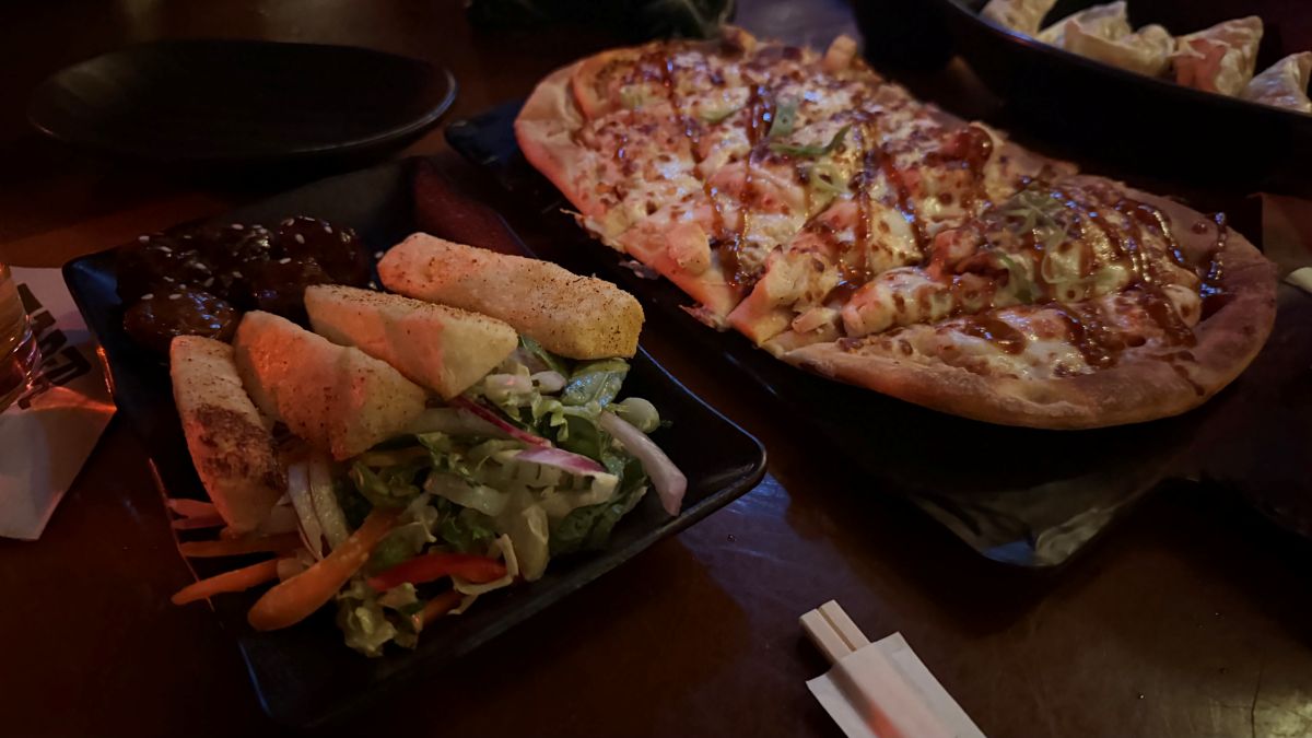 A flatbread pizza drizzled with sauce alongside a plate of seasoned potato wedges and a small side salad on a dark restaurant table.