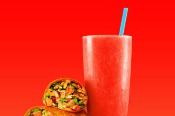A Tiki Torch wrap from Tropical Smoothie Cafe cut in half, stacked next to a tall glass of red smoothie with a bright blue straw, all set against a vibrant solid red background.