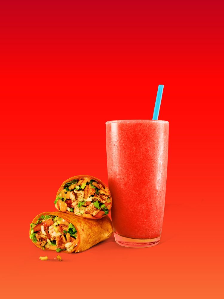 A Tiki Torch wrap from Tropical Smoothie Cafe cut in half, stacked next to a tall glass of red smoothie with a bright blue straw, all set against a vibrant solid red background.