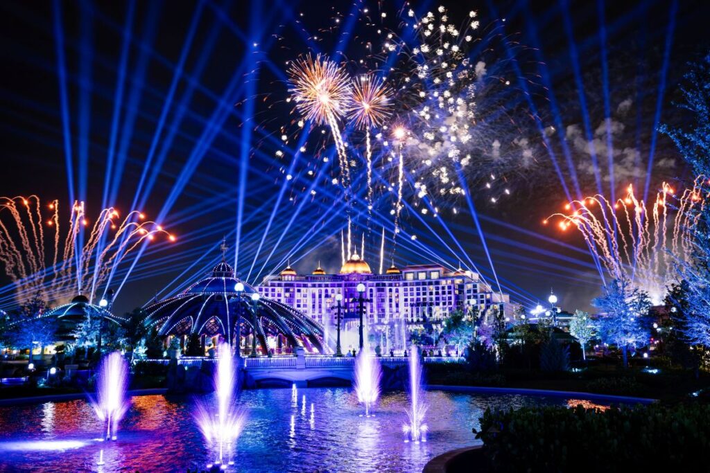 Nighttime view of Universal Epic Universe featuring a large golden firework exploding over a grand resort hotel, with illuminated fountains and a domed structure lit in vibrant red and blue.