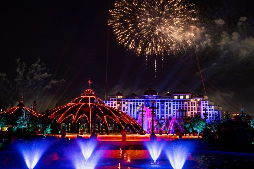 A nighttime spectacular at Universal Epic Universe with multiple fireworks bursting and bright blue laser beams filling the sky above a grand hotel and illuminated water fountains.