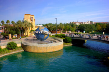 The iconic Universal Studios rotating globe sits at the entrance of Universal Orlando Resort, next to a stone bridge over a clear blue waterway under a sunny sky, serving as a promotion for the Universal Florida Resident Ticket Deal.