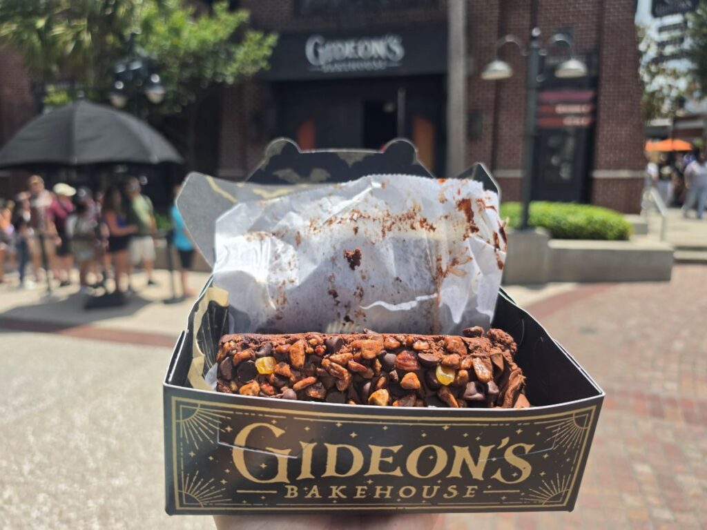 A hand holding an open black Gideon's Bakehouse box containing a thick slice of chocolate cake coated in chocolate chips, pecans, and candied orange pieces.