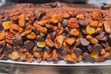 A macro close-up showing the textured side of an orange pecan chocolate cake from Gideon's Bakehouse, completely covered in chocolate chips, chopped pecans, and translucent candied orange bits.