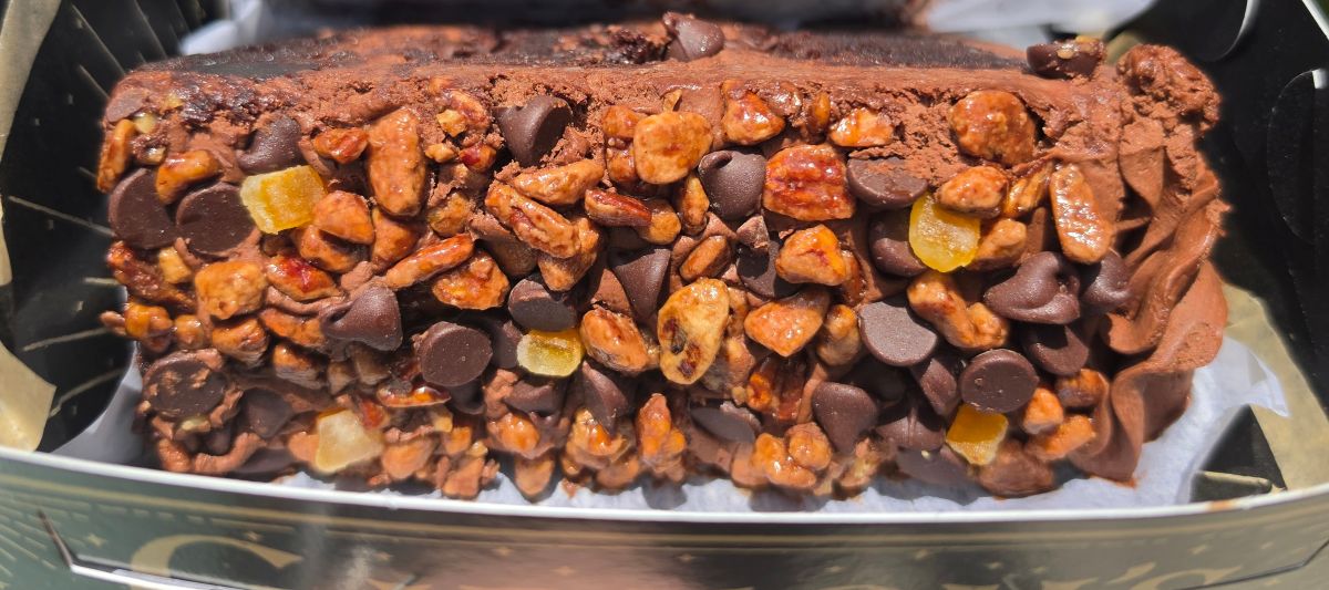 A macro close-up showing the textured side of an orange pecan chocolate cake from Gideon's Bakehouse, completely covered in chocolate chips, chopped pecans, and translucent candied orange bits.