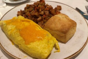 A hearty breakfast plate containing a folded egg omelet topped with melted cheddar cheese, a side of seasoned roasted breakfast potatoes, and a square, golden-brown biscuit.