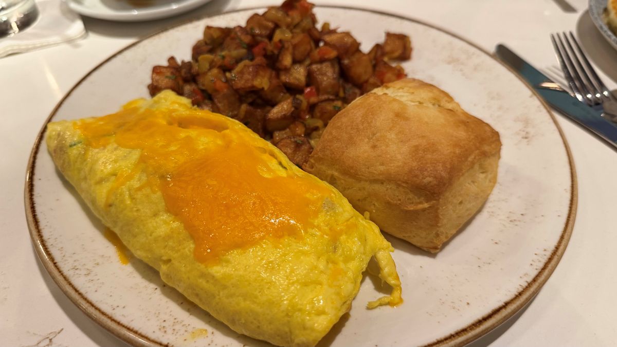 A hearty breakfast plate containing a folded egg omelet topped with melted cheddar cheese, a side of seasoned roasted breakfast potatoes, and a square, golden-brown biscuit.