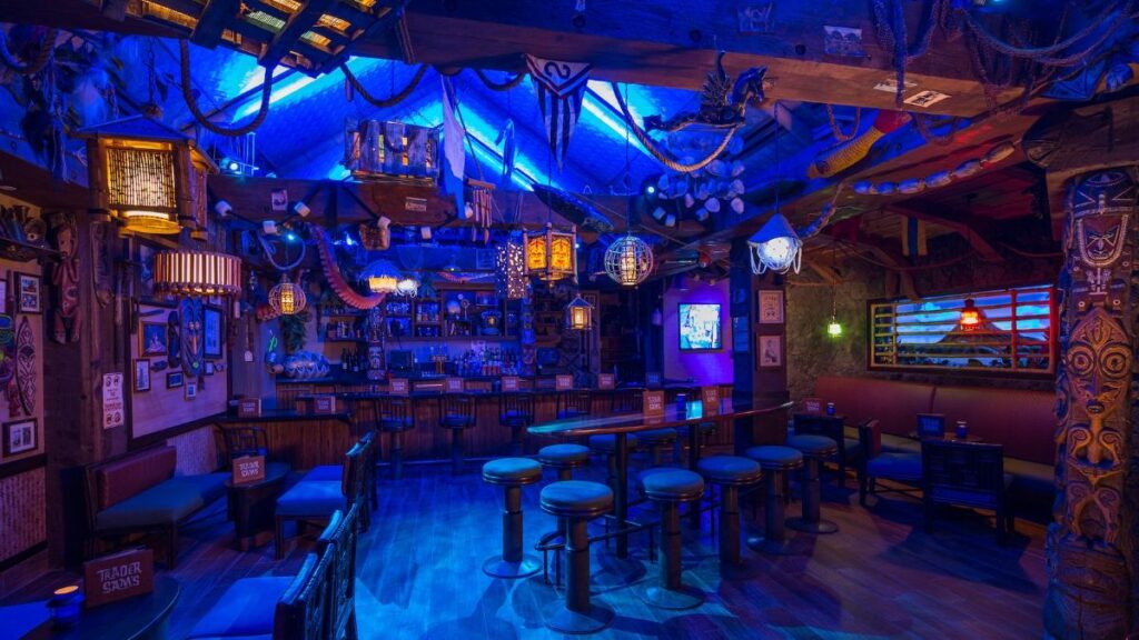 The interior of Trader Sam's Grog Grotto bar bathed in deep blue atmospheric lighting, featuring tiki decor, bar stools, and hanging lanterns.