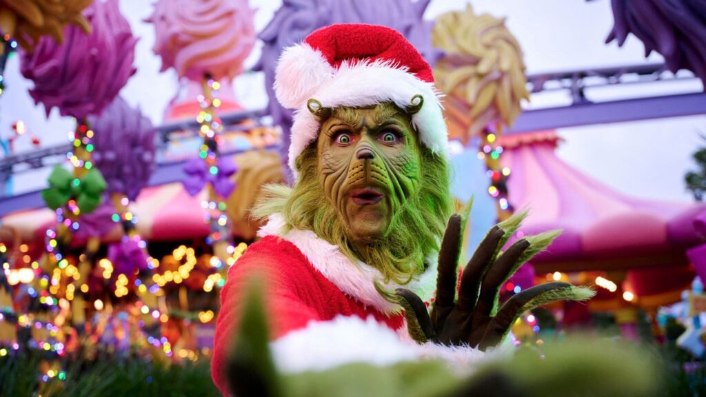 The Grinch dressed in a Santa suit reaching toward the camera with Christmas lights in the background