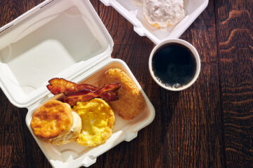 7-Eleven back-to-school deals - A breakfast meal in a white takeout container with a biscuit, egg, bacon, and hash brown, next to a cup of black coffee on a dark wooden table.