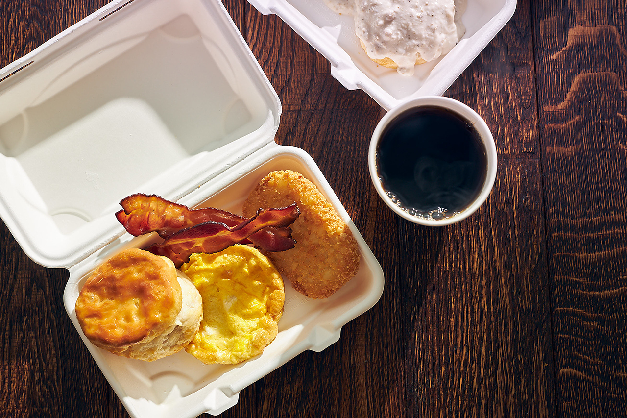 7-Eleven back-to-school deals - A breakfast meal in a white takeout container with a biscuit, egg, bacon, and hash brown, next to a cup of black coffee on a dark wooden table.