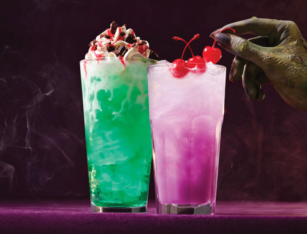 Applebee's Halloween mocktails including the green Graveyard Soda with whipped cream and cookie crumbles next to a purple Tropical Spell Mocktail garnished with cherries.