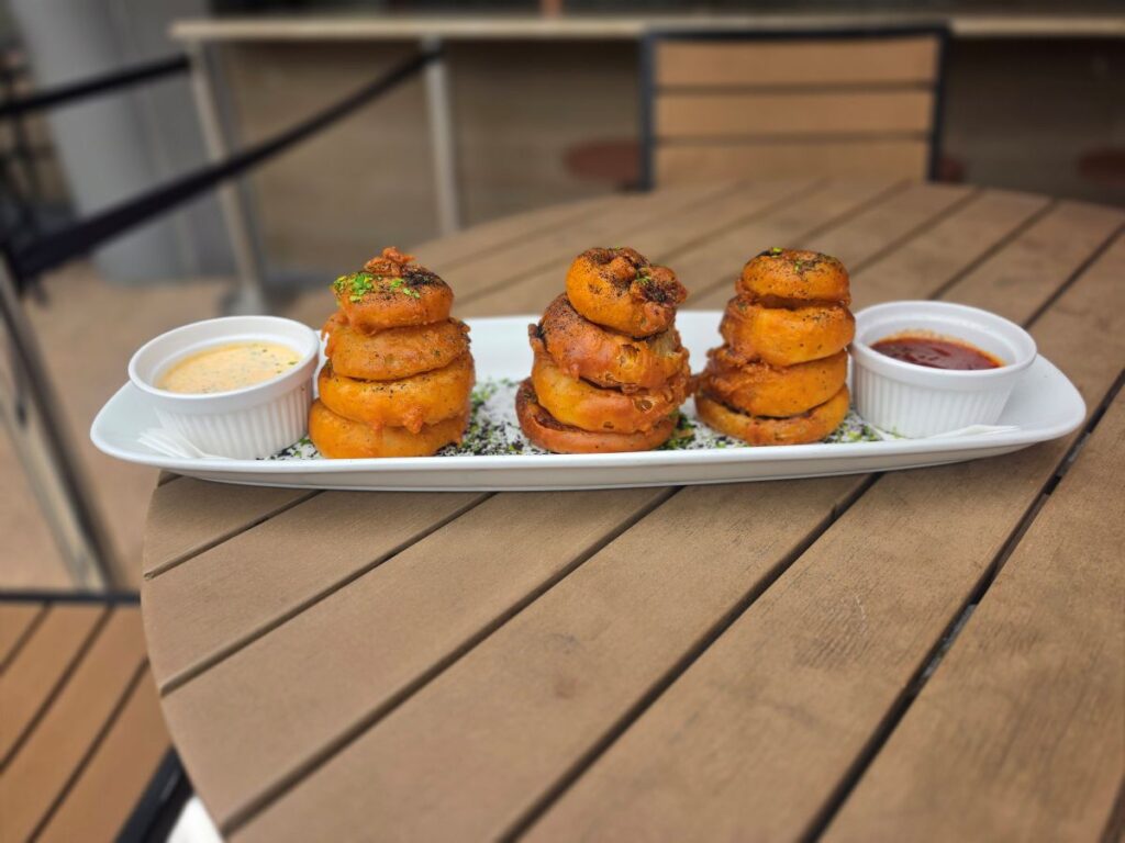 Beer battered onion rings stacked on a white plate with dipping sauces on an outdoor wooden table/