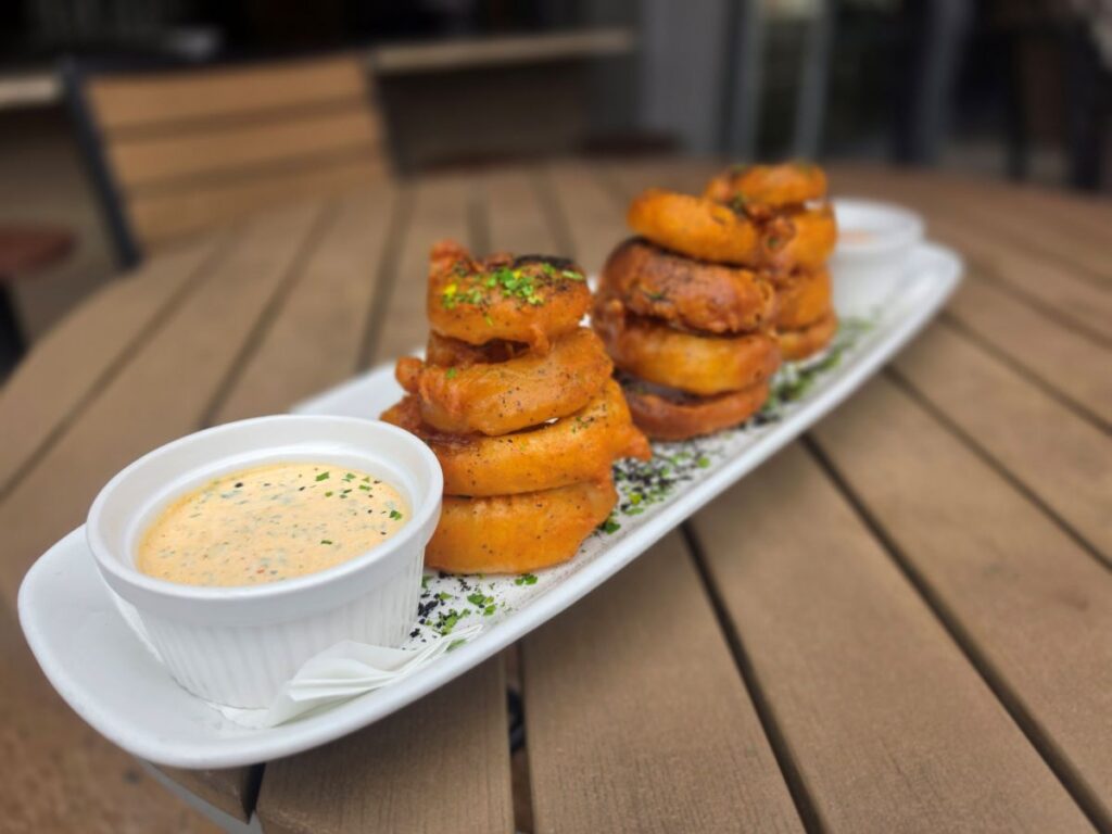 Close-up of beer-battered onion rings stacked on a white dish with dipping sauce for City Works Chef Selects menu.