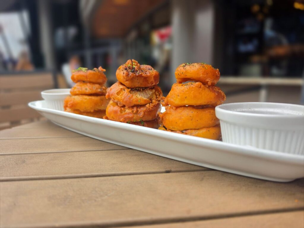 Beer battered onion rings stacked on a white plate with dipping sauces on an outdoor wooden table at City Works.