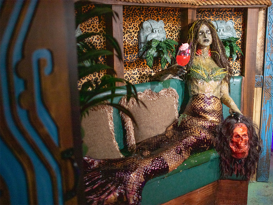 Horrific monster mermaid with scales and seaweed holding tropical drink and severed head