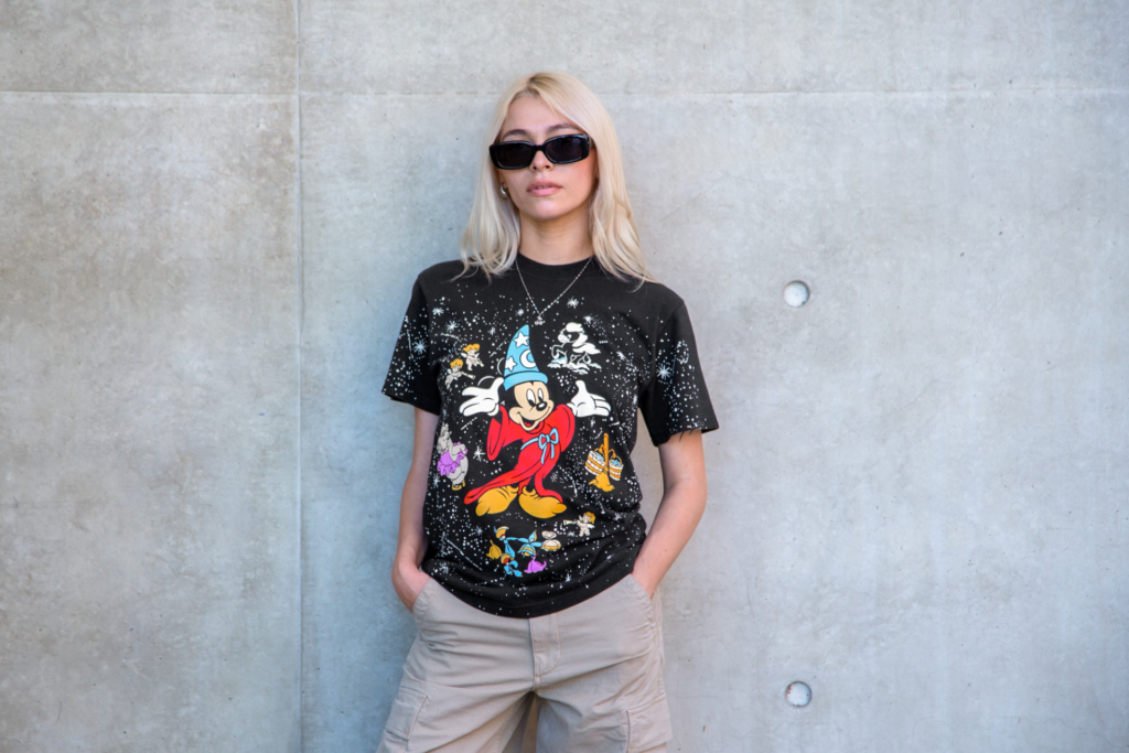A blonde woman wearing black sunglasses and a black t-shirt featuring Mickey Mouse as a sorcerer stands against a plain grey concrete wall background.