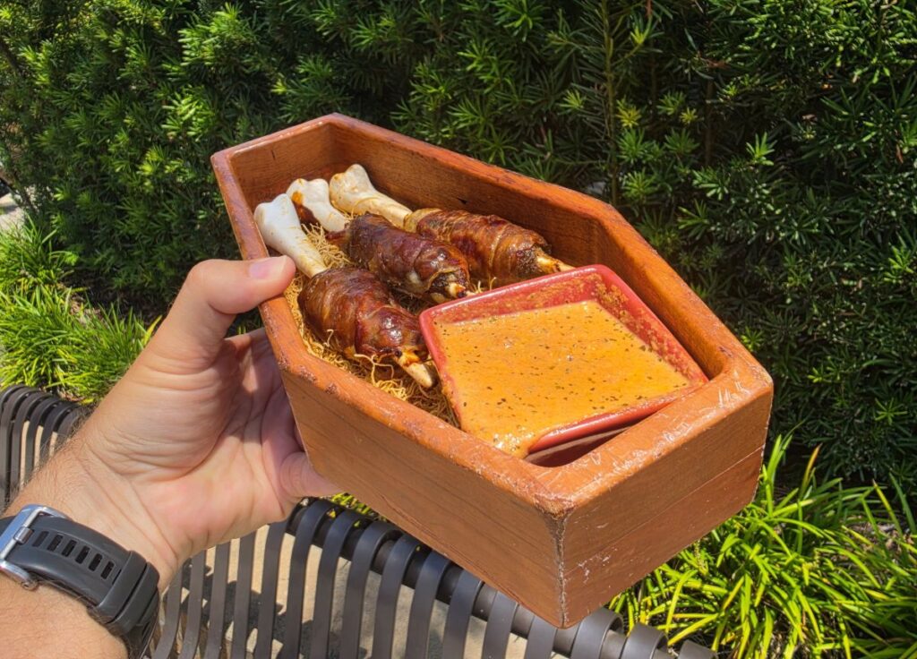 Three prosciutto-wrapped pork and beef bone kebabs served in a coffin-shaped wooden vessel alongside creamy tomato sauce at Jock Lindsey's Hangar Bar.