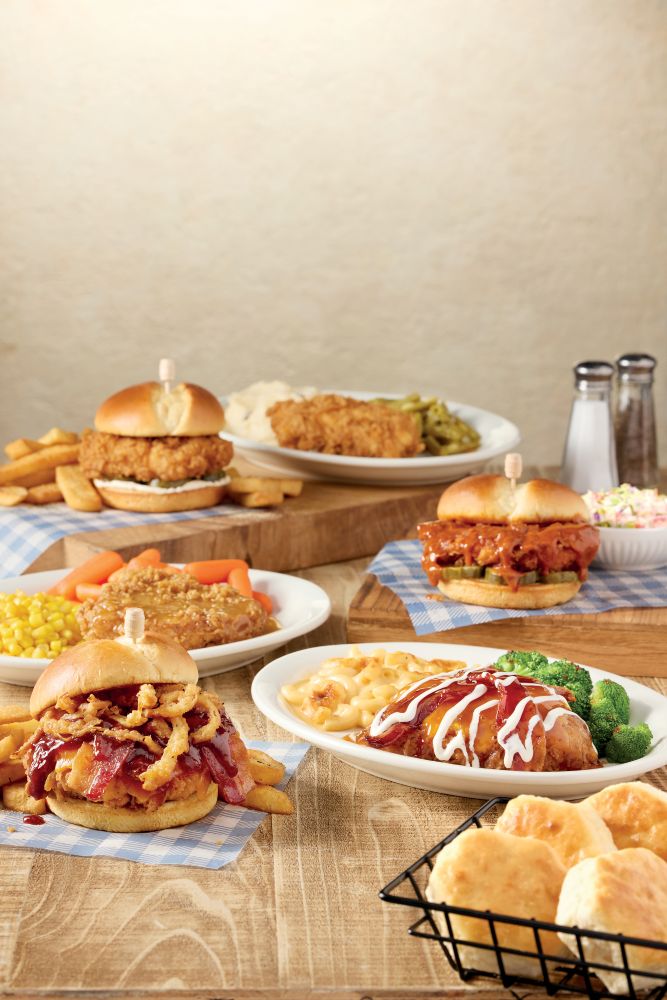 Assortment of Cracker Barrel fried chicken sandwiches, homestyle chicken entrees with sides, and a basket of freshly baked biscuits arranged on a wooden table.