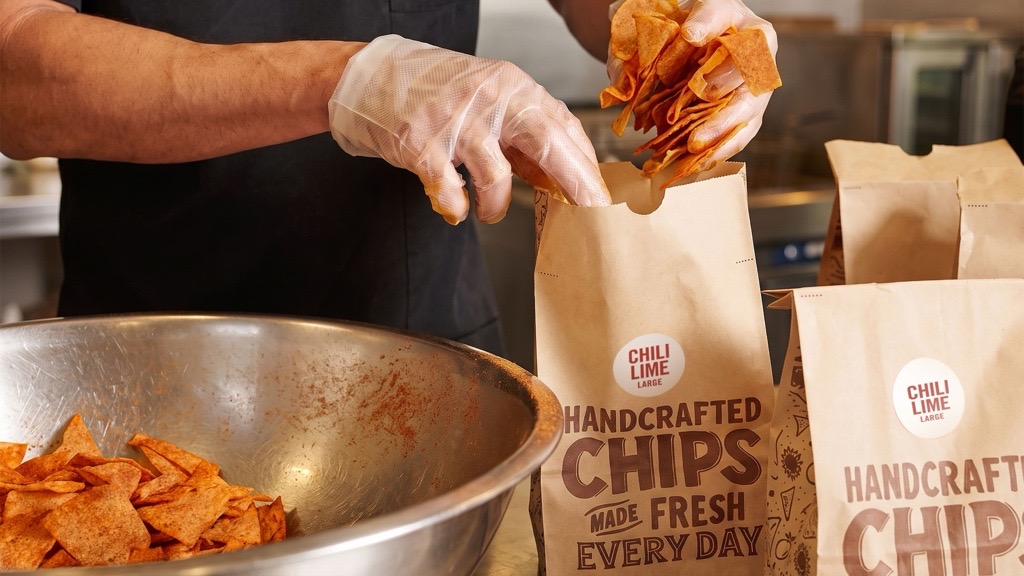 A worker wearing gloved hands scooping seasoned Mexican tortilla chips into a brown paper bag labeled Handcrafted Chips.