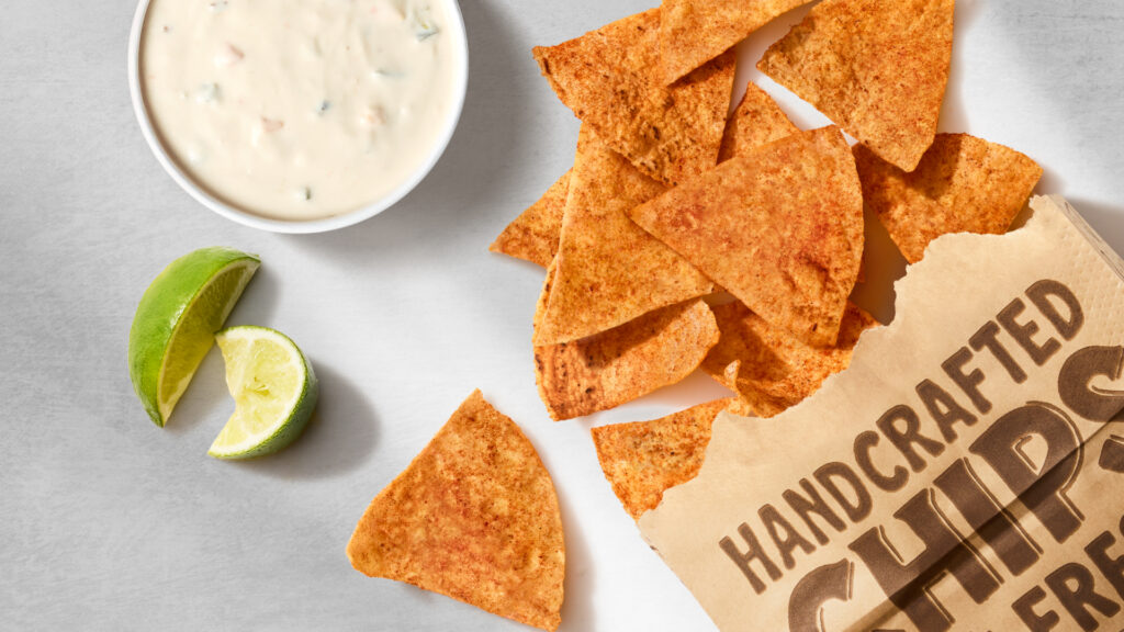 Crispy tortilla chips seasoned with chili lime seasoning spill out of a Chipotle paper bag next to fresh lime wedges and a side dish of queso blanco.
