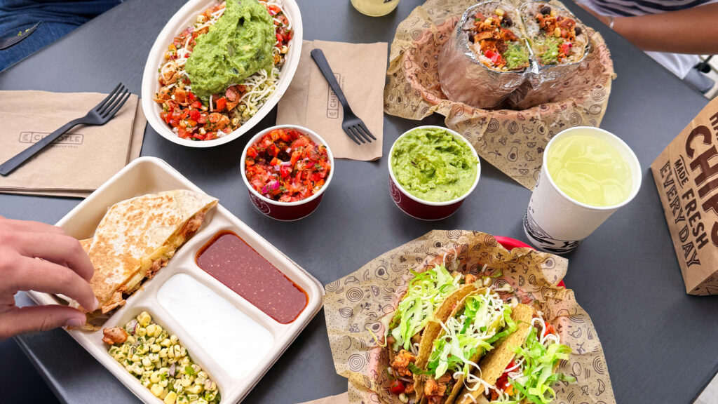 A top-down view of a Chipotle meal featuring a burrito bowl, tacos, a quesadilla with sides, salsa, guacamole, and a drink on a dark table surface.