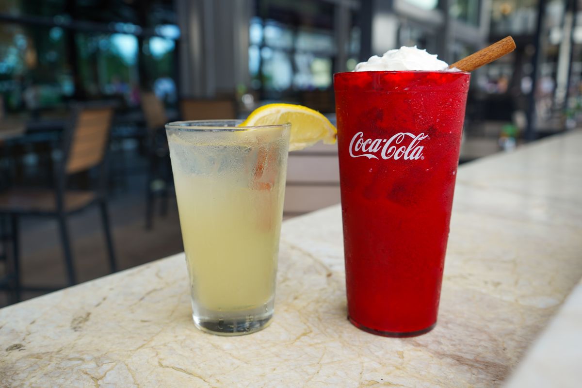A glass of lemonade with a lemon wedge next to a red Coca-Cola cup topped with whipped cream and a cinnamon stick on a marble bar counter surface for City Works Pumpkin Spice beverages promotion