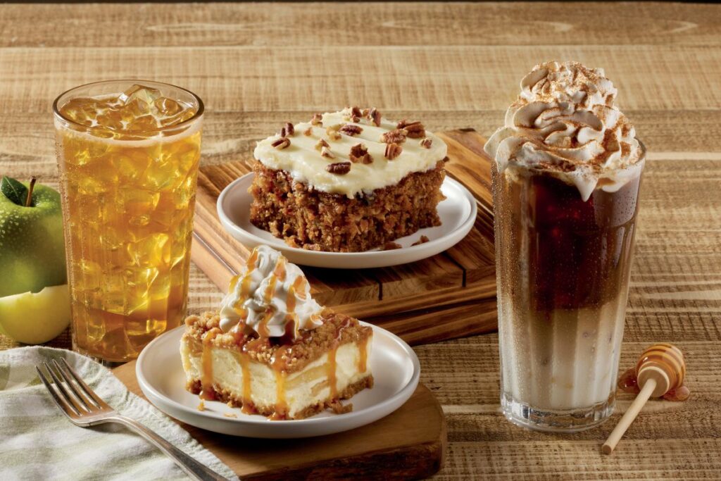 Slice of carrot cake topped with pecans, caramel apple cheesecake drizzled with sauce, an iced apple drink, and an iced latte topped with whipped cream and cinnamon.