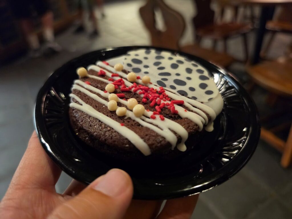 A hand holds a black plate bearing a round Pinocchio Village Haus MNSSHP Cruella Cookie Sandwich decorated with white icing, small white candy spheres, and red sprinkles in a dimly lit room.