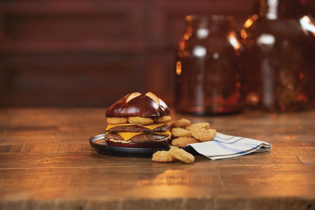 Culver's Back to School Menu: A double cheeseburger on a pretzel bun topped with fried pickles, served with a side of tater tots on a black plate on a rustic wooden table.