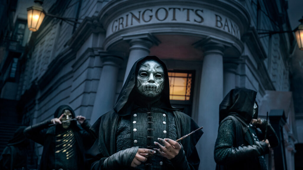 Three figures in dark hooded cloaks and silver masks stand before the Gringotts Bank building, holding wands in a dimly lit, atmospheric setting.