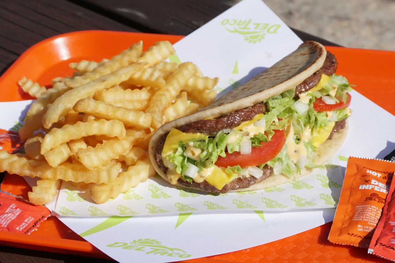 A Del Taco Burgertaco on a pita with lettuce, tomato, cheese, and sauce, served with crinkle-cut fries on an orange tray with hot sauce packets nearby.