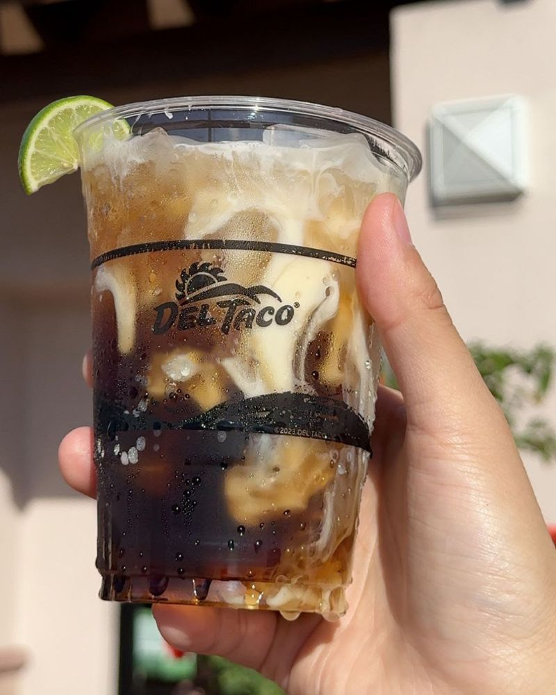 A hand holds a clear plastic Del Taco cup filled with iced coffee and cream, garnished with a fresh lime wedge on the rim against a blurred wall.