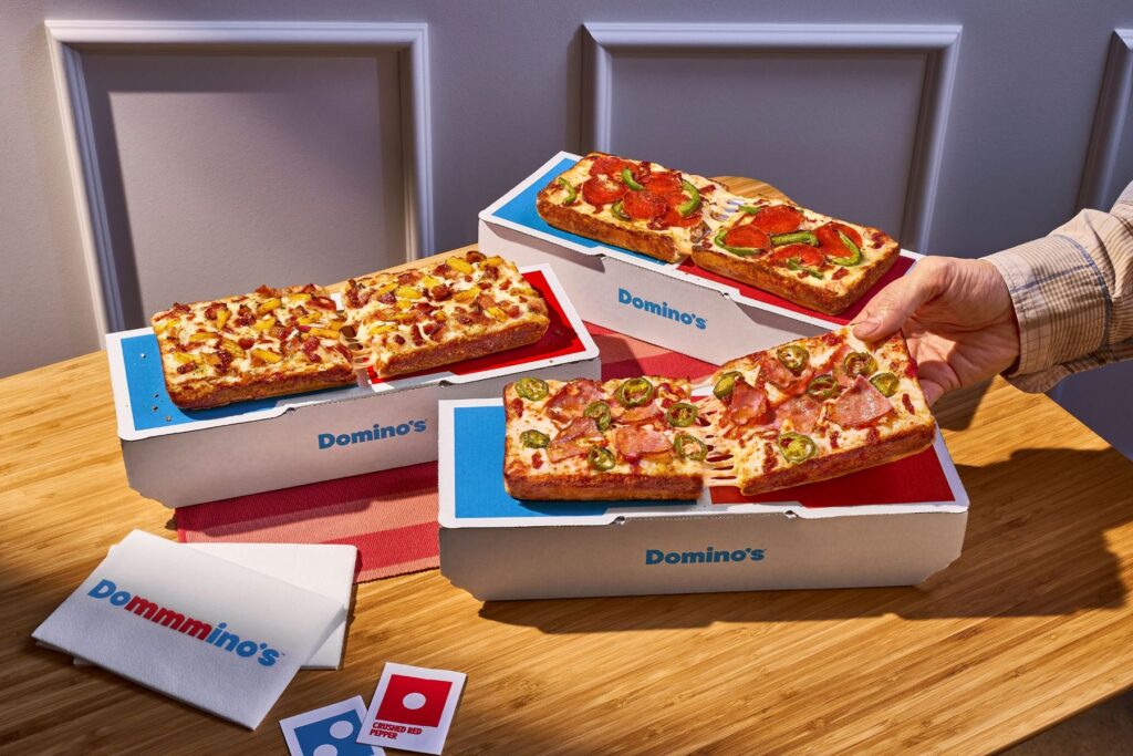 A hand pulls a slice of rectangular Domino's pizza with ham and jalapeños from a box, with other pizza boxes and branded napkins on a wooden table.