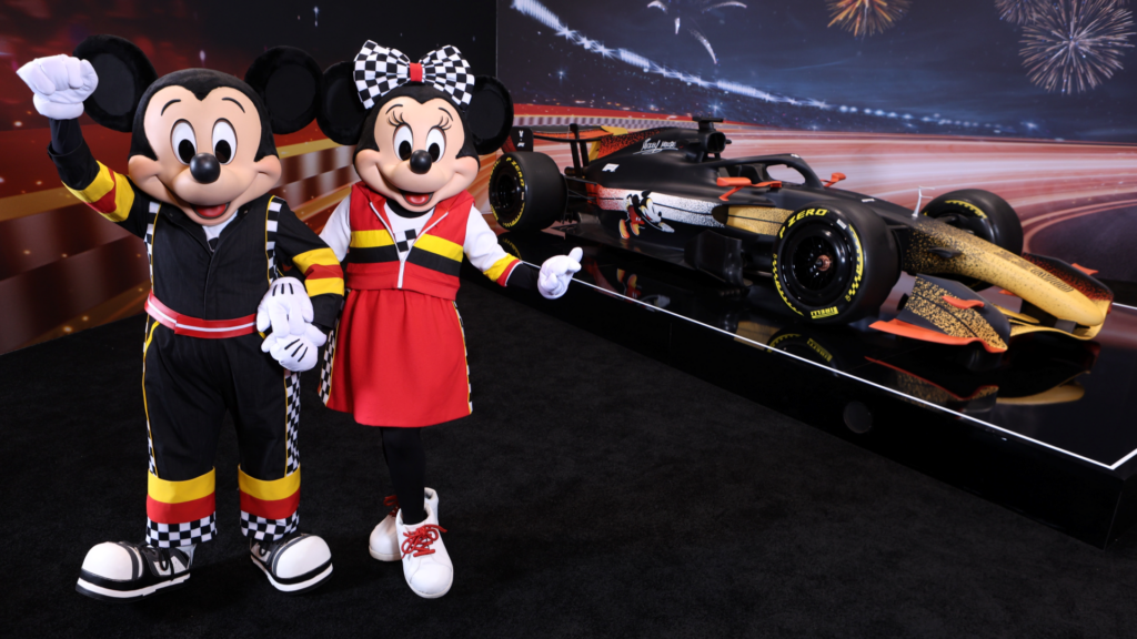 Mickey Mouse and Minnie Mouse in racing suits posing next to a custom black and gold Disney Formula 1 Cars.