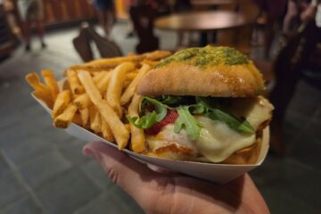 A hand holds a paper tray with a Fra Diavolo chicken sandwich on a pesto-topped bun, melted cheese, arugula, and a side of golden-brown crispy French fries.