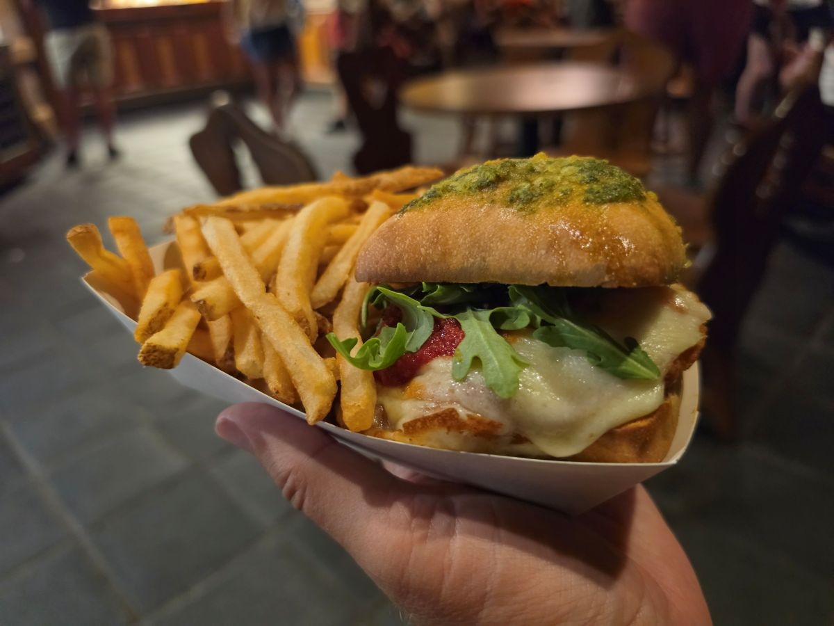 A hand holds a paper tray with a Fra Diavolo chicken sandwich on a pesto-topped bun, melted cheese, arugula, and a side of golden-brown crispy French fries.