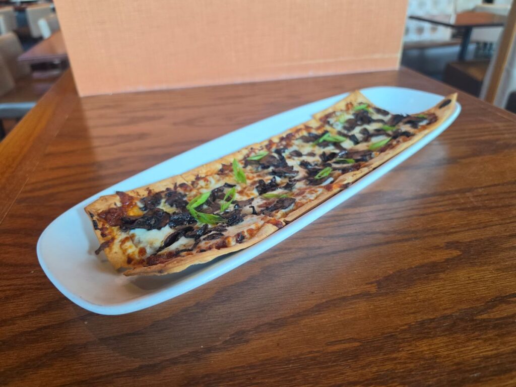 Long white serving platter holding a sliced French onion flatbread on a wooden table for City Works Chef Selects menu.