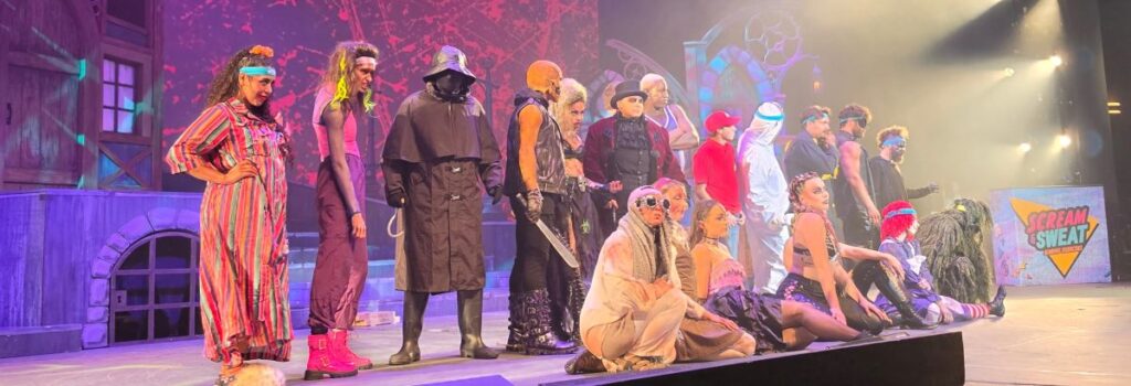 A group of performers in diverse costumes stands on a stage with a red backdrop, featuring a sign that reads Scream & Sweat in a retro font style.