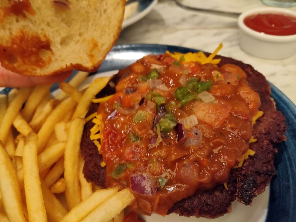 An open Impossible Chili Burger showing a thick layer of chili with diced tomatoes, peppers, and melted cheese topped with french fries on the side.