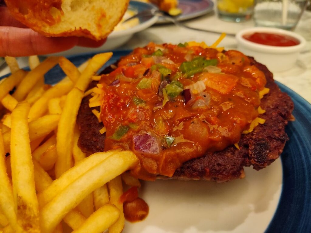 A close-up view of a Chili Impossible Burger with the top bun lifted to display the chili topping and fries on a plate.