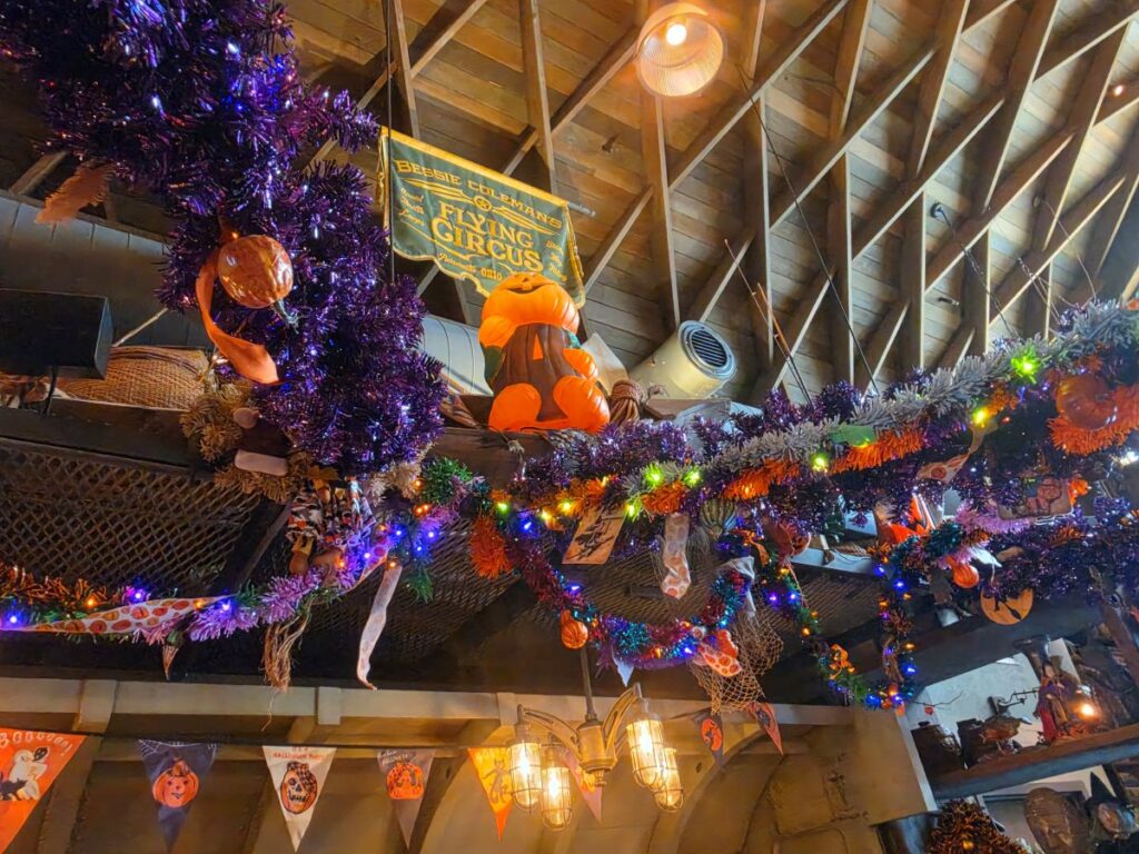 Halloween decorations featuring purple tinsel, orange pumpkins, light strands, and thematic banners hanging from the ceiling rafters inside Jock Lindsey's Hangar Bar.