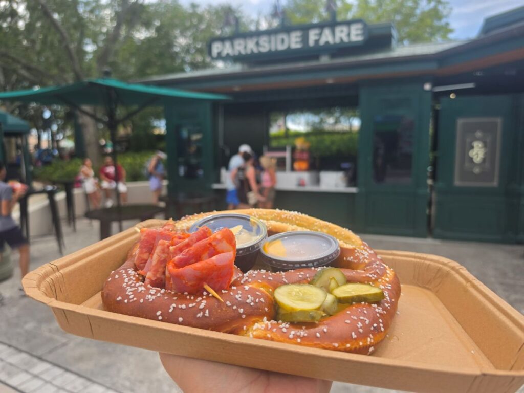Close-up of a salted Loaded Bavarian Pretzel with cured meats on a skewer, pickle slices, and sauce cups outside Parkside Fare at Universal Studios Florida.