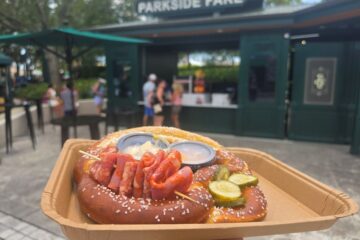Loaded Bavarian Pretzel topped with sliced meats, pickles, and dipping sauces on a cardboard tray in front of Parkside Fare at Universal Studios Florida.