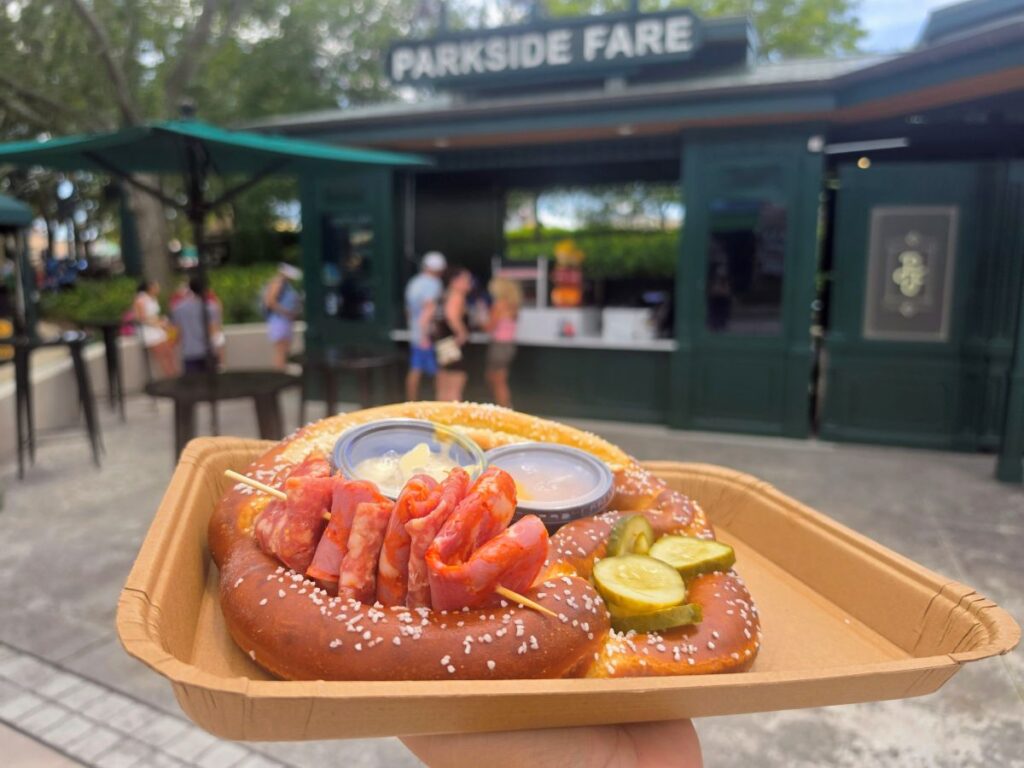 Side view of a Large Pretzel served with meats, pickle slices, and dipping sauces held in front of the Parkside Fare dining location at Universal Studios Florida.