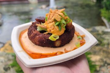A close-up view of a Loaded Brisket Burnt End Stuffed Potato Ball with chipotle lime crema, beer cheese, crispy onions, and sliced scallions, presented in a square paper dish held over a blurred outdoor setting at Busch Gardens Bier Fest